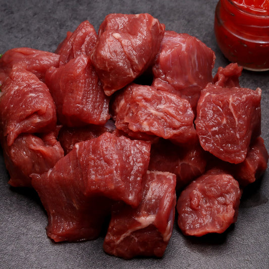 Raw beef chunks on a dark surface with a small container of red sauce and green herb.